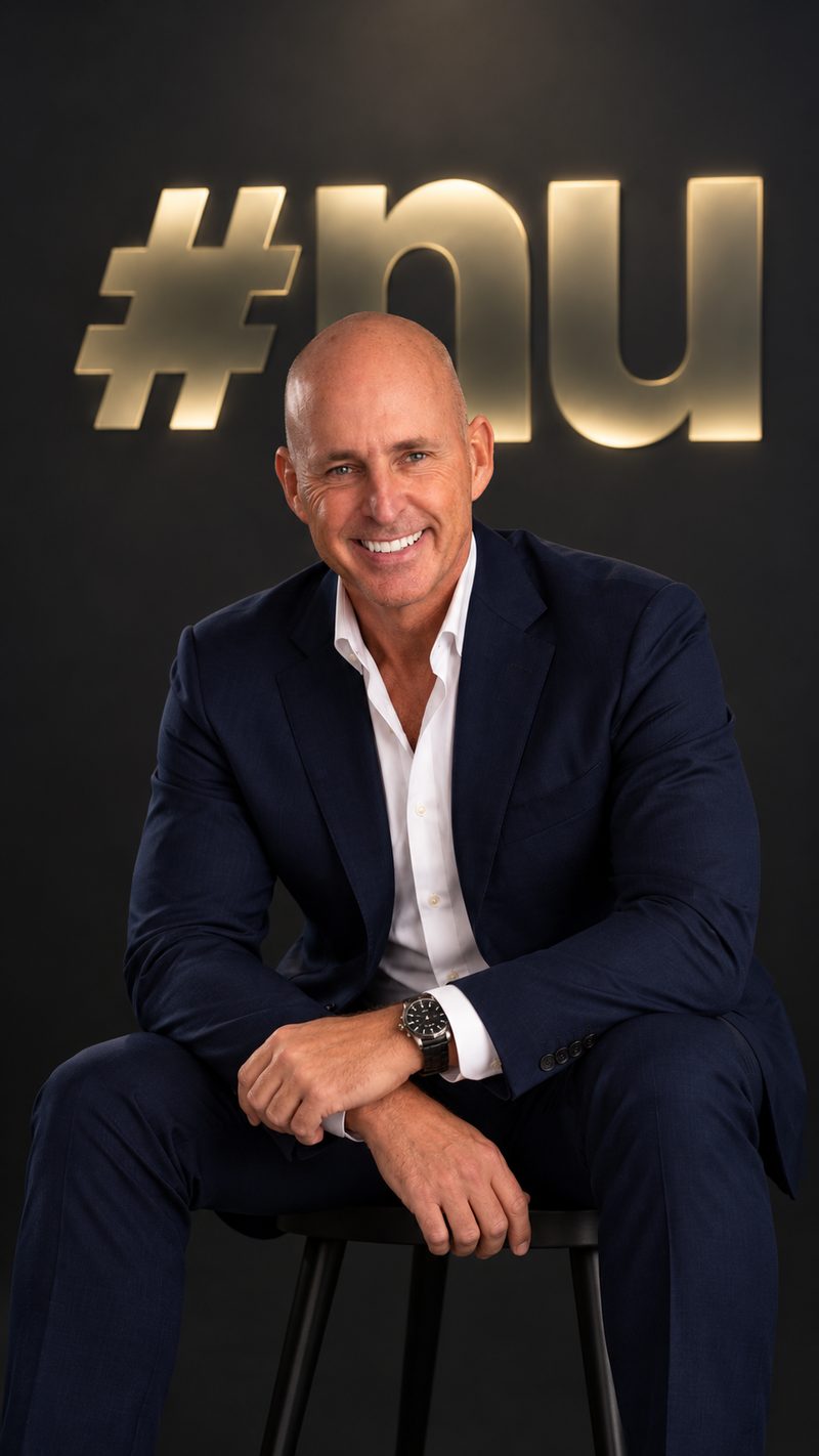 Nick Jeffries, founder of NU Projects, smiling, seated on a stool in a navy suit below the gold #nu sign