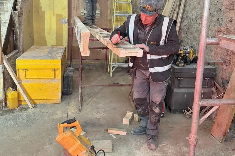 A carpenter on site marking out a length of timber with a square and pencil.